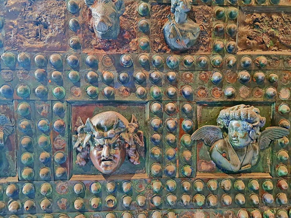 VII.2.18 Pompeii. October 2023. 
Arca/strong box, detail from front of box – 
(on left) a mask with long hair crowned with ivy, and (on right) the head of a winged cupid richly crowned with grape leaves.
Photo courtesy of Giuseppe Ciaramella. 
