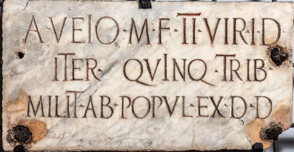HGW02 Pompeii. On the 16th March 1763 a marble plaque was found set in the tufa block on the back of the schola. 
According to Mau the marble plaque had the inscription

A VEIO M F IIVIR I D
ITER QVINQ TRIB
MILIT AB POPUL EX D D

Now in Naples Archaeological Museum. Inventory number 3887.

According to Epigraphik-Datenbank Clauss/Slaby (See www.manfredclauss.de) this read
 
A(ulo) Veio M(arci) f(ilio) IIvir(o) i(ure) d(icundo)
iter(um) quinq(uennali) trib(uno)
milit(um) ab(!) popul(o) ex d(ecreto) d(ecurionum)      [CIL X 996]

According to Cooley this translates as 

To Aulus Veius, son of Marcus, duumvir with judicial power twice, quinquennial, military tribune by popular demand. By decree of the town councillors.

See Kockel V., 1983. Die Grabbauten vor dem Herkulaner Tor in Pompeji. Mainz: von Zabern. (p. 51).
See Cooley, A. and M.G.L., 2004. Pompeii: A Sourcebook. London: Routledge. (p. 139, G4).

