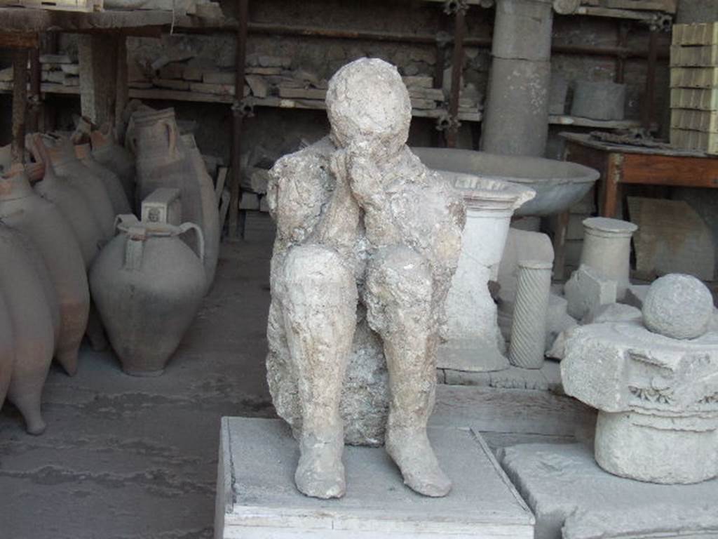 Victim 28.  On display in VII.7.29 Pompeii. May 2006. Plaster cast of body and items in storage.
Two victims were found towards the eastern extremity from the southern portico, and from which plaster-casts could be made because they both lay in the high layer of ash. The one was a beautiful young man's body with agile legs knocked down on the portico floor at the first fall of the ashes.

The other, the most tragically posed, was found leaning against the wall, kneeling, crouching on the ground, in that crouching position that is found in Neolithic tombs, with his head folded forward, bent almost between his knees, for the better protection of himself with his hands and cloak against the inhalation of asphyxiating gases. He had shoes on his feet and wore the typical cloak of the peasant, the “cuculla”, whose flaps he held tight against his mouth.  And in removing the ash under his knees, imprints of overlapping fabrics were noticed, as if the unfortunate man had found comfort in his tragic pose by shielding his knees with the few garments he could arrange. (p.225-6)

For Maiuri’s “Scavo della “Grande Palestra” nel quartiere dell’Anfiteatro, - see Notizie degli Scavi, Anno 1939, Fascicoli 7, 8, 9, on pages 165-238.

