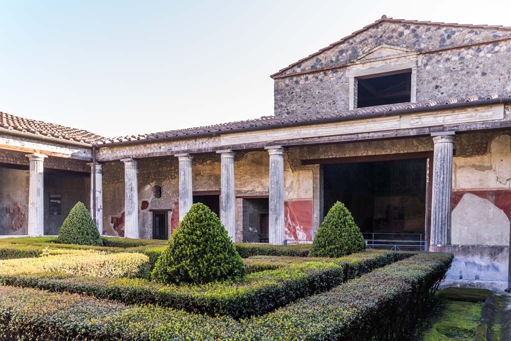 I.10.4 Pompeii. April 2022. 
Looking across peristyle garden towards east side from south-west portico. 
The pediment and window above room 18 can be seen. Photo courtesy of Johannes Eber.

