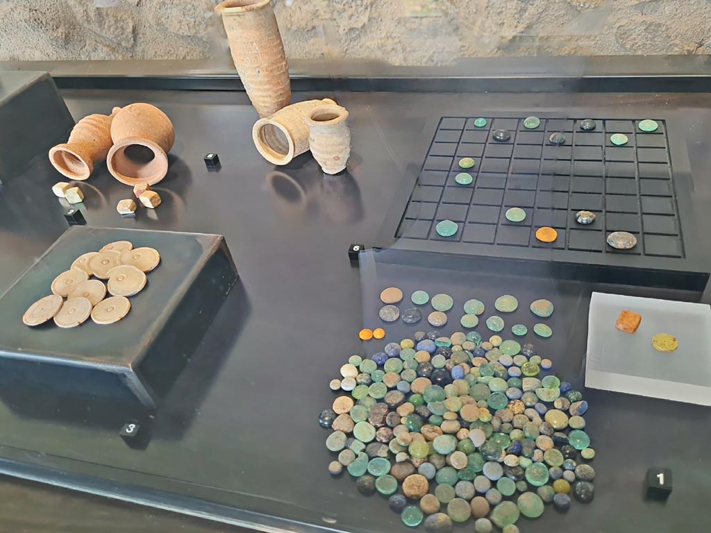 II.1.6 Pompeii. March 2024. No.5 above, in upper centre of photo, pottery dice shakers. Photo courtesy of Giuseppe Ciaramella.
On display in exhibition in Palaestra entitled – “L’altra Pompei, vite comuni all’ombra del Vesuvio”. 
No.1 – Gaming counters made of glass paste, die and tesserae made of bone, from Porta Stabia Necropolis.
No.2 – Worked bone dice, from V.4.13, Pompeii.
No.3 – Gaming counters made of worked bone, from VI.16.26/27, Pompeii.
No.5 – Pottery dice shakers, from II.1.6, Pompeii.
No.6 – Reproduction of a gaming table. 
