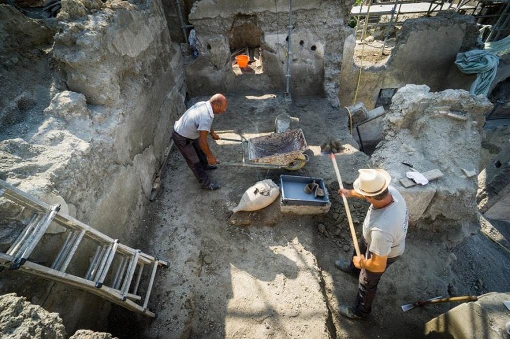 V.2.Pompeii. Casa di Orione. June 2018. 
Looking south across room A3 on the south side of entrance A4, under excavation.
In the upper centre of the photo, the holes for the support beams of the upper floor can be seen, indicating a vaulted alcove at the south end of the room.
When further lapilli/debris was removed from the floor area, a bed recess was seen in the west wall of the room.
Photograph © Parco Archeologico di Pompei.
See Osanna M, 2019. Pompei. Il tempo ritrovato. Le Nuove Scoperte. Milano: Rizzoli (p. 97). 


