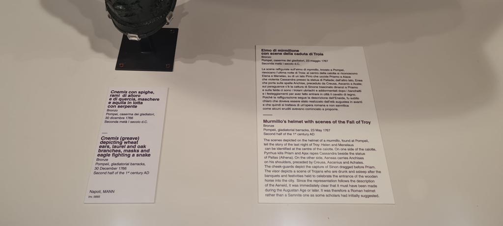 VIII.7.16 Pompeii. Photo taken May 2021, in Naples Archaeological Museum, 
Description card – 
On left - Bronze greave depicting wheat ears, laurel and oak branches, masks and eagle fighting a snake, inv. 5665.
On right – Murmillo gladiator’s helmet with scenes of capture of Troy, discovered Pompeii on 23rd May 1767 in Gladiator’s Barracks, inv. 5673. 
Photo courtesy of Giuseppe Ciaramella.
