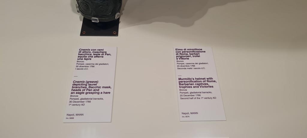 VIII.7.16 Pompeii. Photo taken May 2021, in Naples Archaeological Museum. 
Description card – 
On left - Bronze greave depicting laurel branches, Bacchic masks, heads of Pan and an eagle clasping a hare, inv. 5668.
On right - Murmillo gladiator’s helmet, with personification of Rome, Barbarian captives, trophies and Victories, inv. 5674. 
Found in Gladiator’s Barracks, 20th December 1766. Photo courtesy of Giuseppe Ciaramella.
