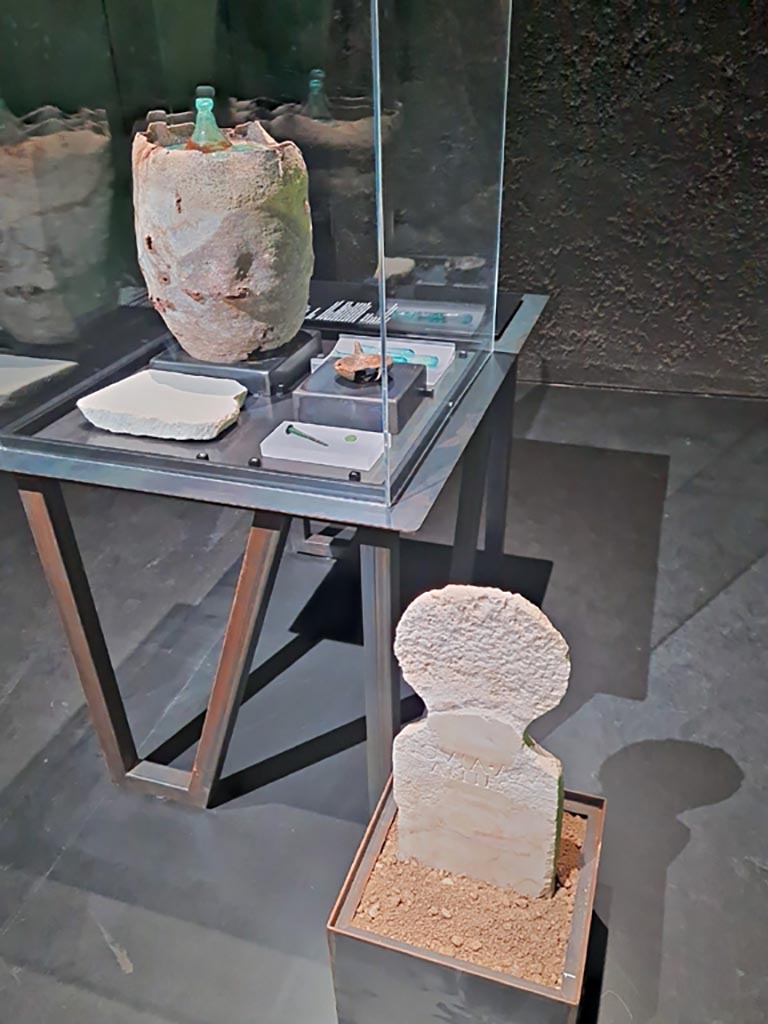 Porta Sarno la tomba di Marcus Venerius Secundio. Tomb of Novia Amabiles. March 2024. Items found in the tomb.
The tomb was found in the burial enclosure of Marcus Venerius Secundus.
Marble columella of Novia Amabiles, presumed to be the wife of Marcus Venerius Secundus.
Her cremated remains were preserved in the glass urn, together with those of two children, most likely the children of the couple.
A terracotta lamp, a bronze nail, a marble slab and glass unguentaria and a bronze coin were also found.
Photo courtesy of Giuseppe Ciaramella.
On display in exhibition in Palaestra entitled – “L’altra Pompei, vite comuni all’ombra del Vesuvio”. March 2024.
