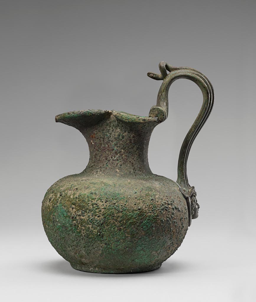 Discovered in a villa in Boscoreale. Bronze oinochoe (pitcher) with snake and gorgon’s head handle.
Digital image courtesy of the Getty's Open Content Program. Now in the Getty Museum, inventory number 72.AC.133.
Except for three concentric incised rings at the join of the neck to the shoulder, the body of this bronze oinochoe (pitcher) is undecorated. At the base of the handle, however, is an attachment in the form of a gorgon’s head. The central rib of the arching handle takes the form of a snake's body, its head modelled free of the handle itself, perhaps for use as a thumb rest. On each side of the handle is a spool-shaped ornament.
Vessels of this type were used in the home for religious rituals and to hold water for handwashing during banquets. Slaves would pour water from a pitcher over the hands of participants, catching the cascade in a patera (shallow bowl) held underneath. This oinochoe was discovered in a villa at Boscoreale or in the vicinity, although the exact findspot is unknown. 
